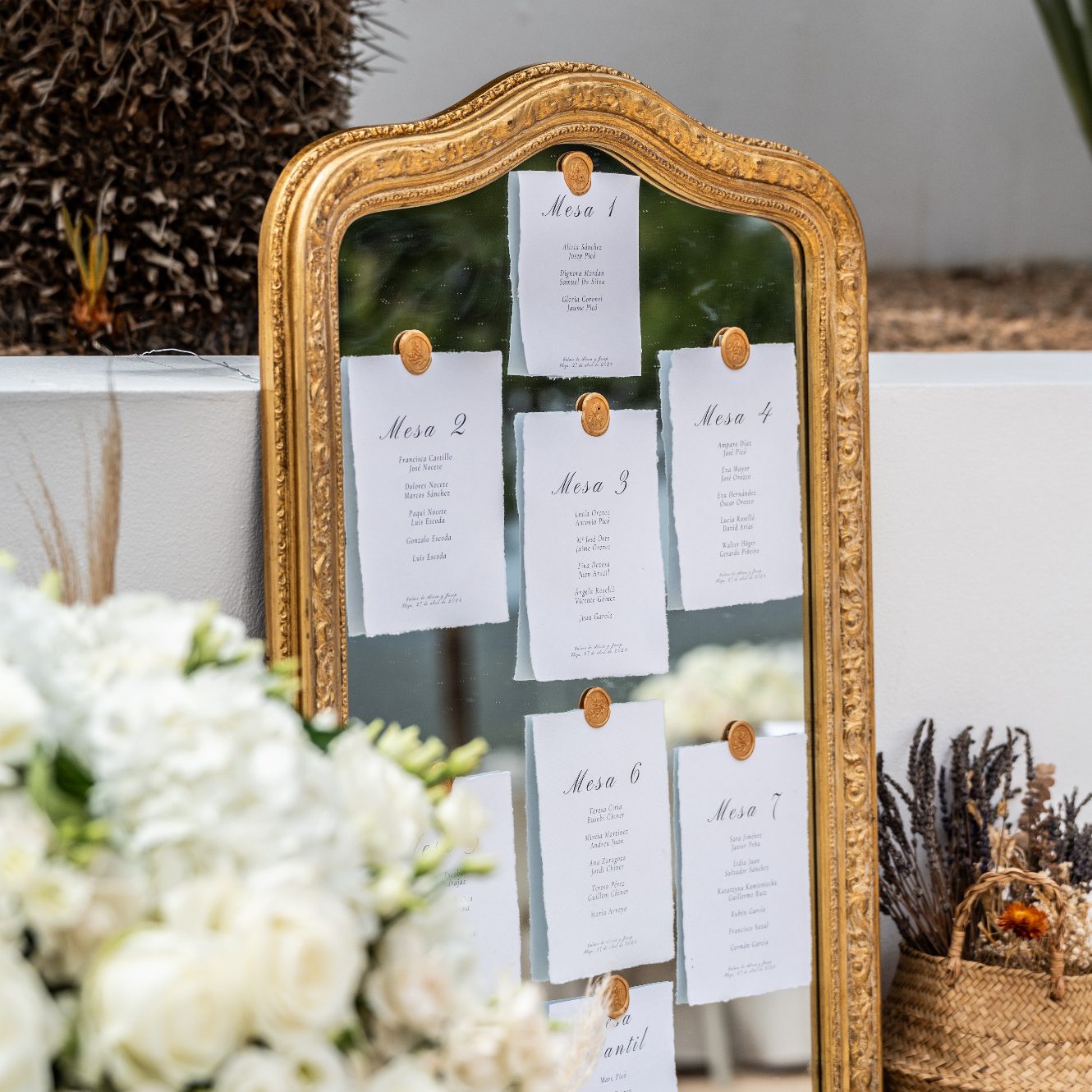 decoración de bodas Espejo dorado con listas de invitados colgadas en tarjetas blancas y flores blancas en el fondo.